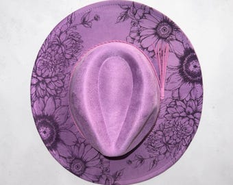 May include: A lavender fedora hat with a floral design. The hat features a lavender crown and brim, with a pink band and tassel. The brim is decorated with a black floral pattern. The hat is a stylish accessory.