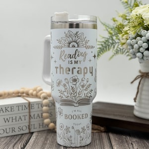 May include: White stainless steel tumbler with a floral design and the text "Reading is my therapy" and "I'm all booked today".