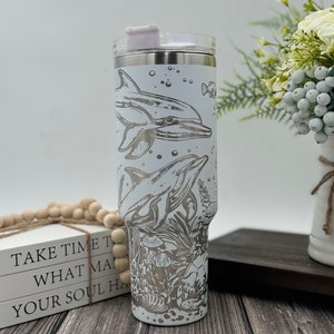 May include: White stainless steel tumbler with a lid and a silver graphic of dolphins and sea life. The tumbler is on a wooden surface.