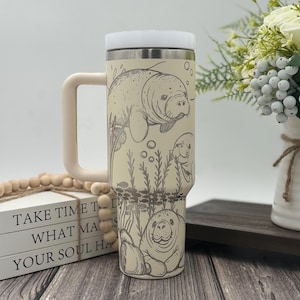 May include: Cream-colored travel mug with a white lid and handle. The mug features an etched design of manatees and aquatic plants. The text "TAKE TIME TO WHAT MATTERS YOUR SOUL HAS" is visible on a stack of books in the background.