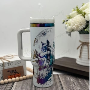 May include: White insulated tumbler with a handle, featuring a wolf design under a full moon. The artwork includes shades of blue, purple, and green. The tumbler has a white lid and a straw.