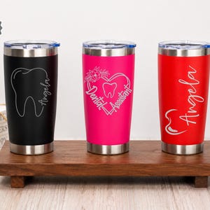 May include: Three stainless steel insulated tumblers in black, hot pink, and red, each with a different dental-themed design and the name "Angela." The black tumbler features a tooth graphic, the pink one a heart, and the red one a cursive name.