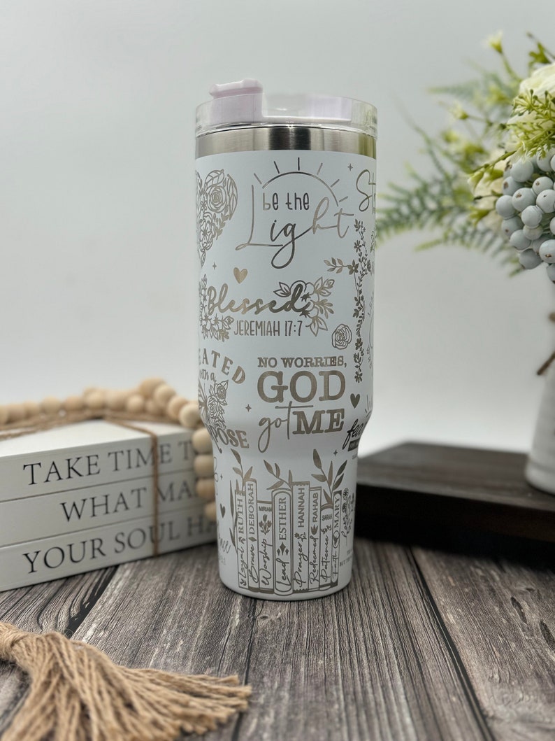 May include: A white stainless steel tumbler with a light pink lid. The tumbler is decorated with floral and heart designs, along with the words "Be the Light", "Blessed", and "God got me". The tumbler also has the text "Jeremiah 17:7".