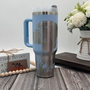 May include: A stainless steel tumbler with a light blue body and handle, featuring a detailed engraved image of a dog. The tumbler has a silver rim and a black lid. The handle is light blue. The tumbler is designed for beverages.