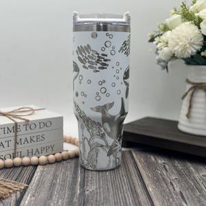 Op de afbeelding: Een witte roestvrijstalen beker met een doorzichtig deksel, met een gegraveerd ontwerp van orka's, scholen vissen en bellen. De beker heeft een zilveren rand en een handvat. Het ontwerp is in grijstinten.