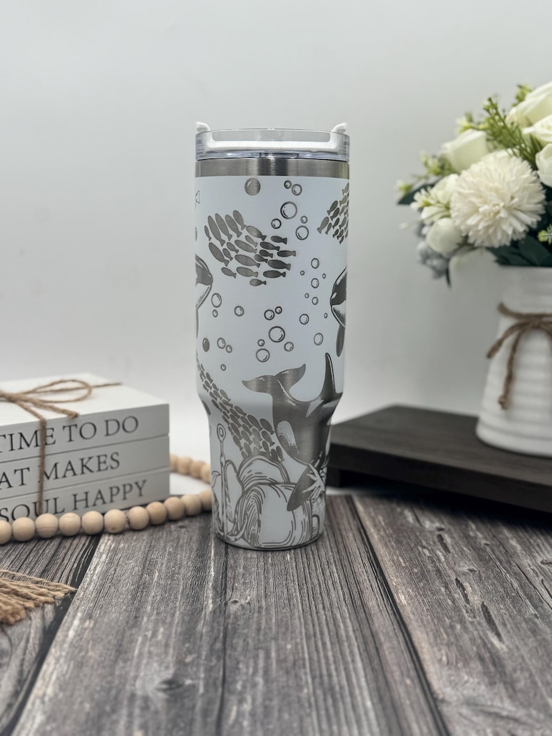 Op de afbeelding: Een witte roestvrijstalen beker met een doorzichtig deksel, met een gegraveerd ontwerp van orka's, scholen vissen en bellen. De beker heeft een zilveren rand en een handvat. Het ontwerp is in grijstinten.