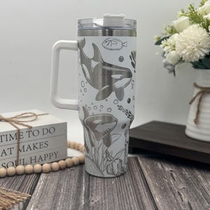 Op de afbeelding: Witte reismok met een handvat, met een rondom ontwerp van orka's, vissen en waterplanten. De mok heeft een doorzichtig deksel en een wit handvat. De tekst op de zijkant luidt "TIME TO DO WHAT MAKES SOUL HAPPY."