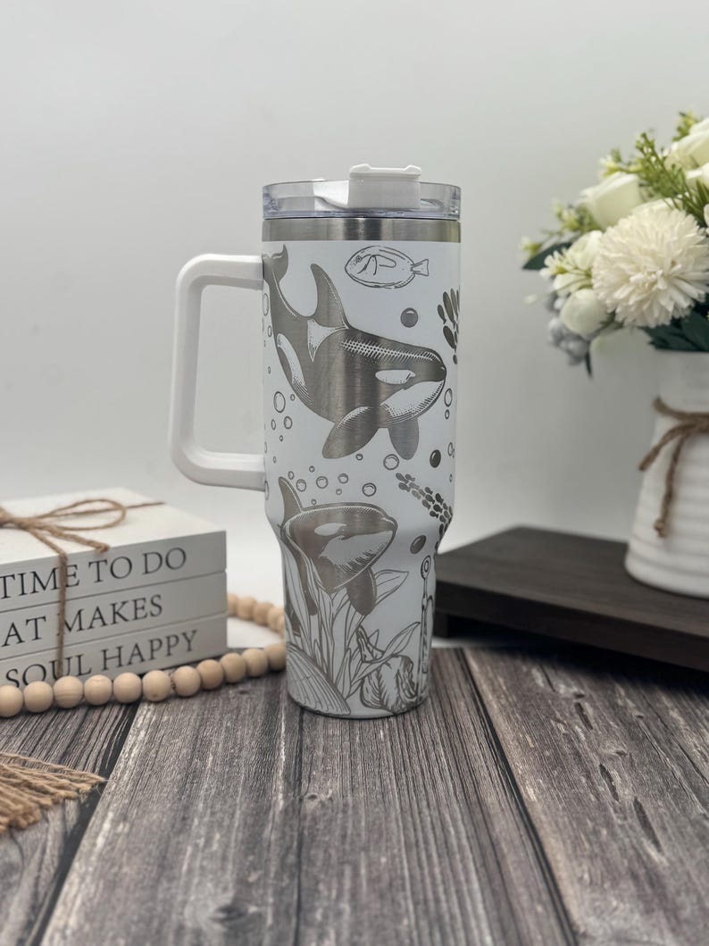 Op de afbeelding: Witte reismok met een handvat, met een rondom ontwerp van orka's, vissen en waterplanten. De mok heeft een doorzichtig deksel en een wit handvat. De tekst op de zijkant luidt "TIME TO DO WHAT MAKES SOUL HAPPY."