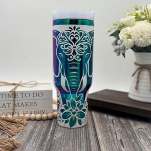 May include: A white stainless steel tumbler with a teal, purple, and blue iridescent finish. The tumbler features a white design of an elephant with intricate floral patterns on its head.