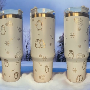 May include: Three white insulated travel mugs with gold penguin and snowflake designs. The mugs have lids and handles.