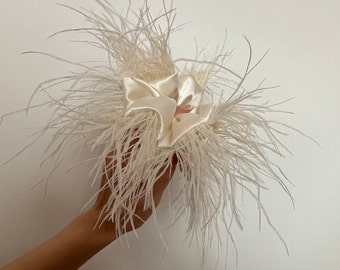 May include: A white feather hair clip with a satin bow in the center. The clip is held in a hand.