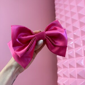 Könnte beinhalten: Eine große, leuchtend rosa Schleife aus Satin mit einer mittleren Schlaufe. Die Schleife wird in einer Hand vor einem rosa Hintergrund gehalten.