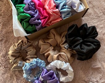 Può includere: Una scatola piena di una varietà di scrunchies di raso colorati. Gli scrunchies sono disposti in file e sono in tonalità di verde, blu, rosa, viola, marrone, nero e bianco.