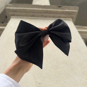 May include: A large black satin bow with a center knot. The bow is held in a hand against a white background.