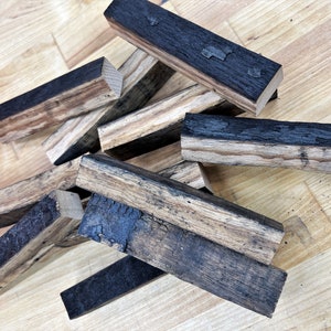 Peut inclure: Un tas de morceaux de bois carbonisés, probablement d'un fût de whisky, de couleur brun foncé et noir. Le bois a une texture rugueuse et est disposé de manière aléatoire.