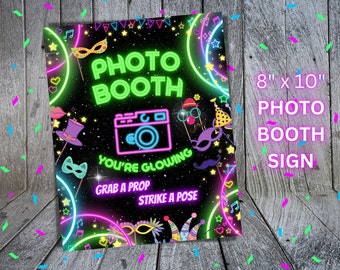 Pode incluir: Um sinal de cabine de fotos verde neon e rosa com fundo preto e estrelas. O sinal diz "Photo Booth" em letras verde neon e "You're glowing" em letras rosa. Também diz "Grab a prop" e "Strike a pose". O sinal mede 8 polegadas por 10 polegadas.