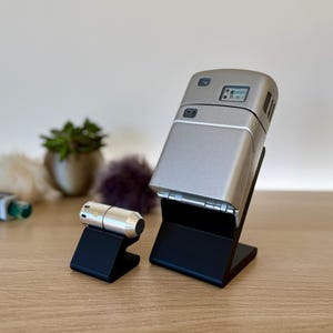 Könnte beinhalten: Zwei silberne elektronische Geräte auf schwarzen Ständern. Das größere Gerät hat ein kleines digitales Display und eine silberne Oberfläche. Das kleinere Gerät ist zylindrisch und hat einen goldenen Akzent.