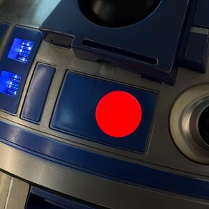 Puede incluir: Primer plano de un robot metálico plateado y azul con un círculo rojo y luces azules. El robot tiene una cámara cilíndrica y paneles rectangulares con detalles azules. El diseño recuerda a un popular personaje de ciencia ficción.