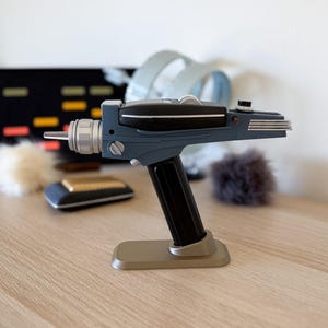 Könnte beinhalten: Eine blaue und schwarze Star Trek Phaser-Replik auf einem goldenen Ständer. Der Phaser hat einen silbernen Lauf und einen schwarzen Griff.