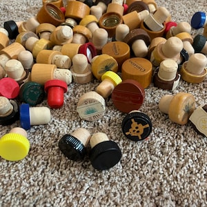 May include: A collection of various bottle stoppers, including cork, plastic and wood. Some stoppers feature designs such as a butterfly, a skull and crossbones, and a star. The stoppers are on a brown carpet.