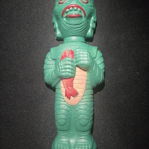 May include: A green monster figurine with red lips and a red object held in its hands. The figurine has a textured, scaly appearance and a light tan midsection. The figure is standing upright on a rounded base.