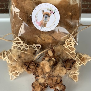 May include: Three brown and white star-shaped ornaments made of woven twigs and filled with alpaca fiber. The ornaments are on a white surface. The image also shows a bag of alpaca fiber with a label that reads "Peaceful Peepas Alpaca Farm & Fiber".