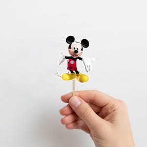 May include: A Mickey Mouse cake topper. The figure is in a standing pose, wearing a red jacket, black shorts, and yellow shoes. The character has white gloves and a smiling expression. The topper is attached to a wooden stick.