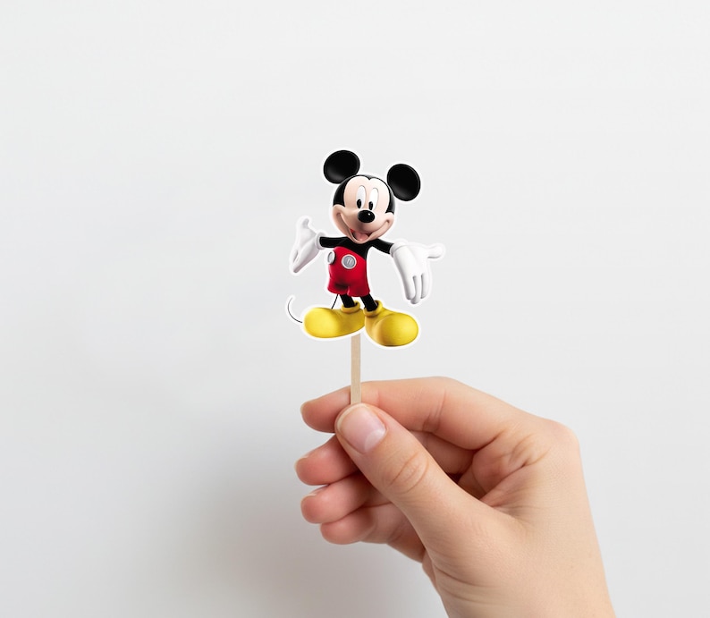 May include: A Mickey Mouse cake topper. The figure is in a standing pose, wearing a red jacket, black shorts, and yellow shoes. The character has white gloves and a smiling expression. The topper is attached to a wooden stick.