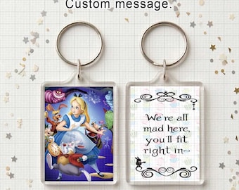 May include: A double-sided rectangular keychain featuring a metal ring. The front side displays a colorful illustration of Alice in Wonderland characters. The back side features a white background with the text We're all mad here, you'll fit right in.