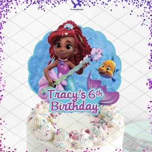 May include: A cake topper featuring a mermaid with red hair, holding a musical instrument, and a yellow fish. The text "Tracy's 6th Birthday" is displayed on the topper. The cake is white with frosting and sprinkles.
