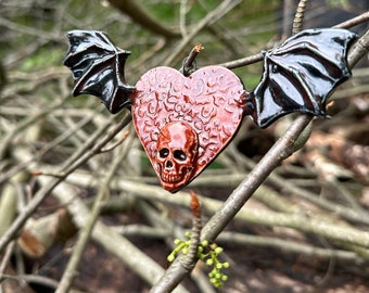 May include: A ceramic heart-shaped pendant with a skull design and black bat wings. The heart is decorated with a floral pattern and the skull is painted in red and white.
