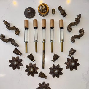 May include: An assortment of antique brass lamp parts, including decorative floral bases, ornate curved arms, and various connectors. The components are arranged on a white surface, showcasing their intricate designs and aged patina.