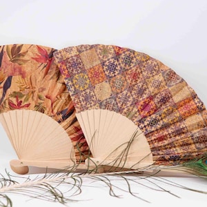 Puede incluir: Dos abanicos decorativos con marcos de madera y papel estampado de colores. Los abanicos están cerrados y de pie sobre una superficie blanca. Una pluma de pavo real está tumbada delante de los abanicos.