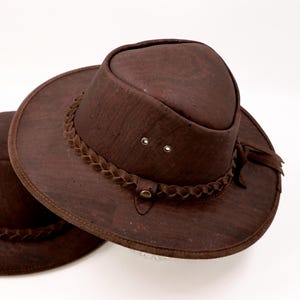 May include: Two brown cork hats with wide brims and braided hatbands. The hats have a classic design with a rounded crown and a decorative band around the base of the crown. The hats are a rich, earthy brown color.