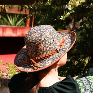 May include: A patterned hat with a brown brim and a decorative leather band. The hat features a repeating blue and beige design. The hat is a stylish accessory for outdoor activities.