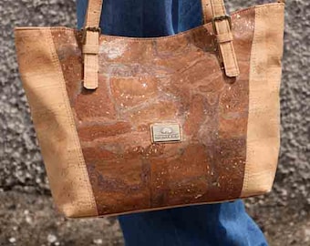 May include: A tote bag made of cork material, featuring a central panel with a marbled effect and lighter cork side panels. The bag has two handles with buckles and a small metal tag. The person is wearing blue jeans and brown shoes.