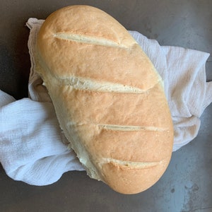 Puede incluir: Una hogaza de pan blanco con una corteza ligeramente dorada, reposando sobre un paño blanco.