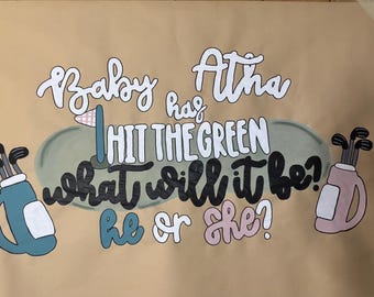 May include: A hand-painted sign on brown paper with the text "Baby Atha has hit the green, what will it be? He or she?" Two golf bags and a golf flag are also illustrated.
