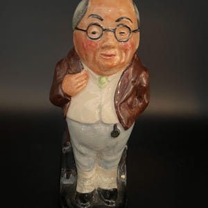 Puede incluir: Una figura de cerámica de una persona mayor calva con gafas redondas, chaqueta marrón, chaleco blanco y pantalones azul claro. La figura tiene una tez sonrosada, una corbata verde y se apoya sobre una base negra con un detalle de reloj de bolsillo.