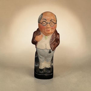 May include: A ceramic figurine of a bald, bespectacled individual with a round face and rosy cheeks. The figure wears a brown jacket over a white waistcoat and light-colored trousers, standing on a black base with a hand placed near the chest.