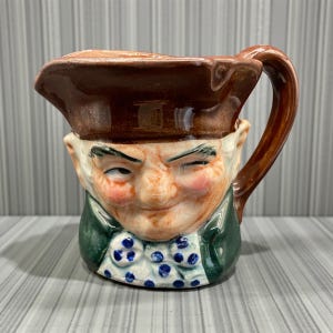 May include: A vintage ceramic pitcher shaped like a man's face, with a brown hat and handle. The face has rosy cheeks, a winking eye, and a green jacket with a blue and white polka dot bow tie. The pitcher is a unique decorative item.