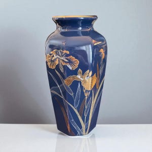 Puede incluir: Un jarrón de cerámica azul marino con diseños de flores de iris doradas. El jarrón tiene forma rectangular con una parte superior ligeramente ensanchada y un borde dorado. Las flores de iris están detalladas con detalles dorados y tallos blancos.