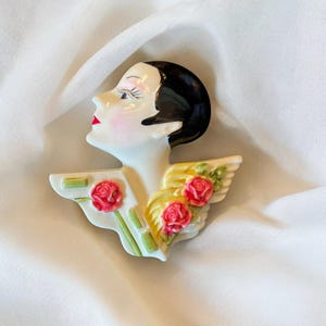 May include: A white ceramic wall plaque featuring a woman's face with black hair and red lips. The woman is wearing a yellow and green floral design around her neck.