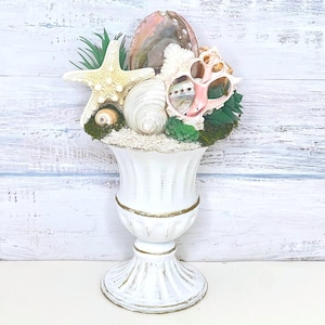 May include: A white pedestal vase with gold-toned trim filled with a collection of seashells, including a large starfish, an iridescent abalone shell, and various spiral shells, accented with green faux succulents and moss on a bed of white sand.