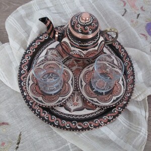Puede incluir: Un juego de té turco tradicional con una bandeja de cobre, una tetera y dos vasos. La bandeja está decorada con intrincados patrones florales en negro y cobre.