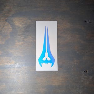 May include: A rectangular sticker featuring a blue and teal energy sword design. The sticker has a gradient effect, transitioning from blue to teal. The design is centered on a white background.