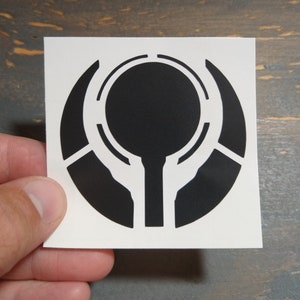 May include: Black and white sticker featuring a stylized symbol with a circle and a stem. The symbol is reminiscent of a futuristic or sci-fi design.