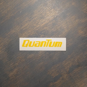 May include: A rectangular sticker with the word "Quantum" in yellow, stylized font. The sticker is on a gray wooden surface. The sticker's font is bold and modern.