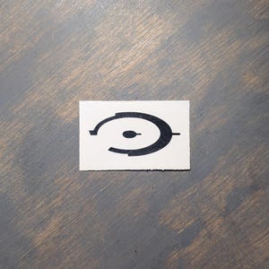 May include: A black and white sticker featuring the Halo video game logo. The logo is a stylized, circular design with a central dot and two curved lines. The sticker is rectangular and sits on a wooden surface.