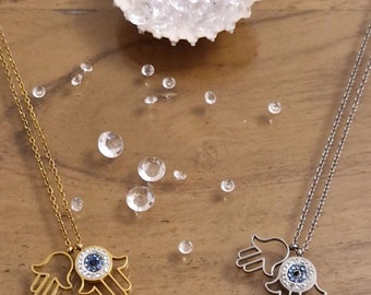 May include: Two gold and silver necklaces with a hamsa hand charm featuring a blue evil eye. The necklaces are on a wooden surface with scattered clear rhinestones.